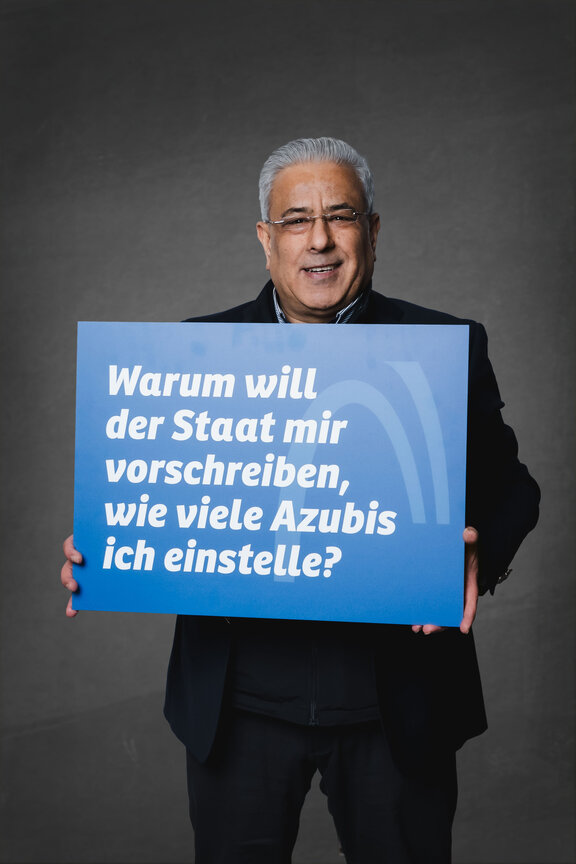 Eine Person steht vor einem neutralen grauen Hintergrund und hält ein großes blaues Schild mit der weißen Aufschrift: "Warum will der Staat mir verschreiben, wie viele Azubis ich einstelle?"