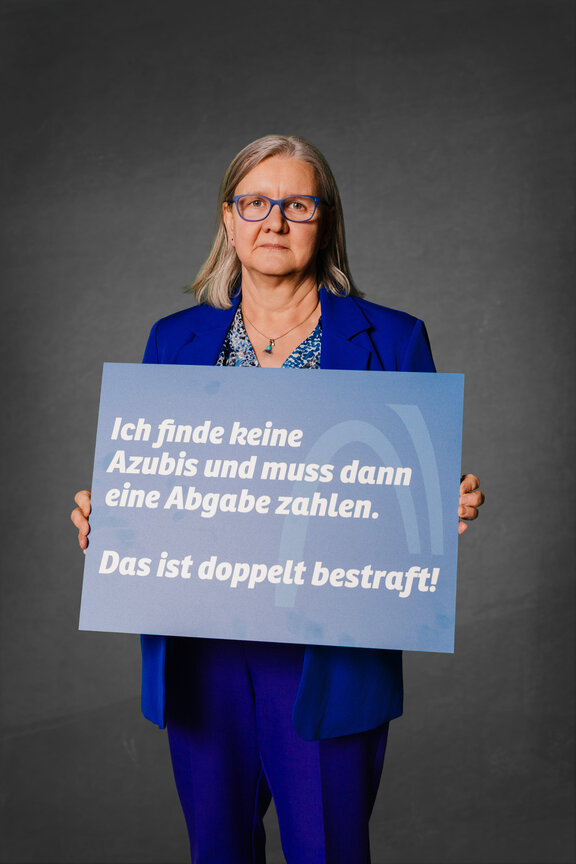 Eine Person steht vor einem neutralen grauen Hintergrund und hält ein großes blaues Schild mit der weißen Aufschrift: "Ich finde keine Azubis und muss dann ein Abgabe zahlen. Das ist doppelt bestraft!"