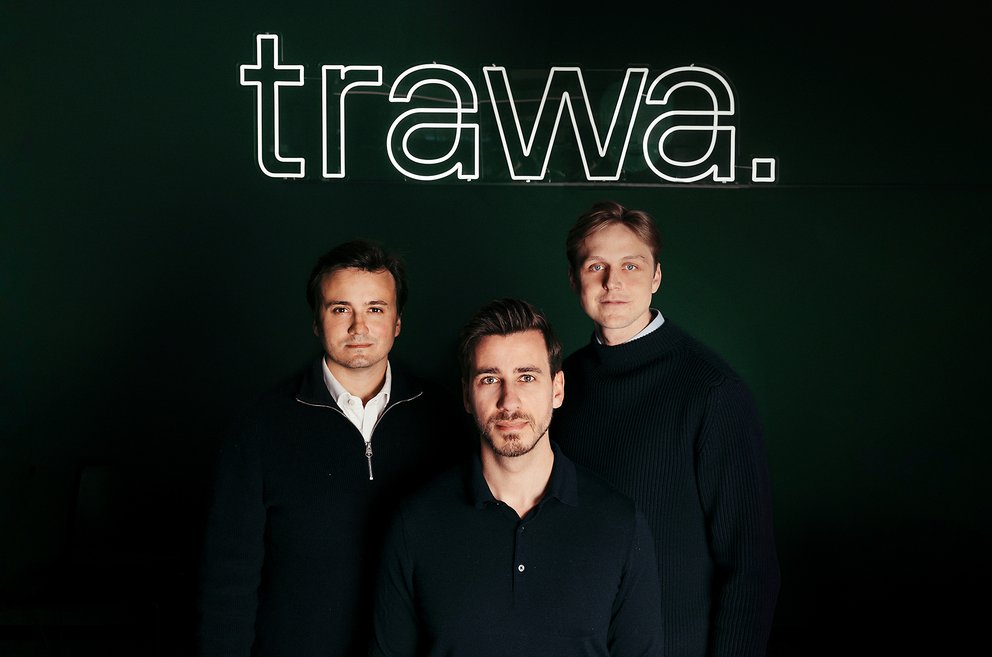 Drei Männer in dunkler Kleidung vor schwarzem Hintergrund mit weiß leuchtendem Schriftzug 'trawa.'