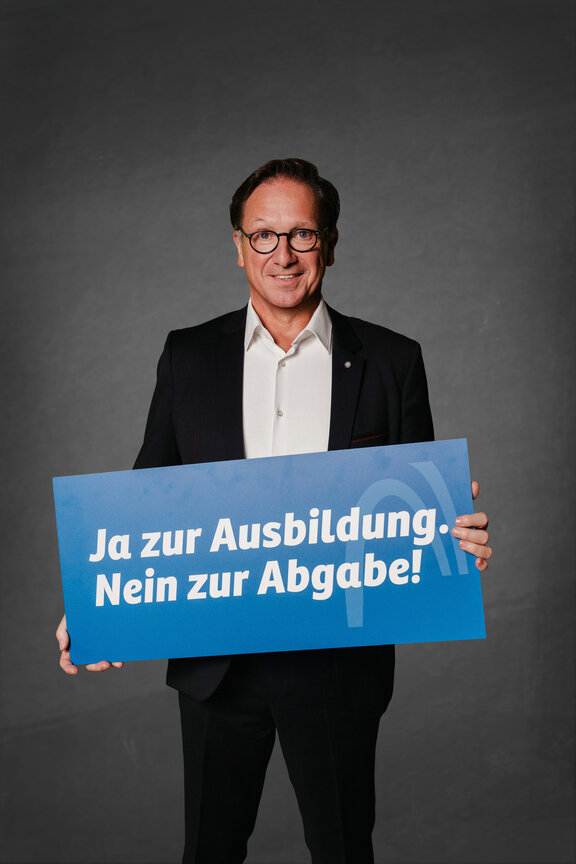 Eine Person steht vor einem neutralen grauen Hintergrund und hält ein großes blaues Schild mit der weißen Aufschrift: "Ja zur Ausbildung. Nein zur Abgabe!"
