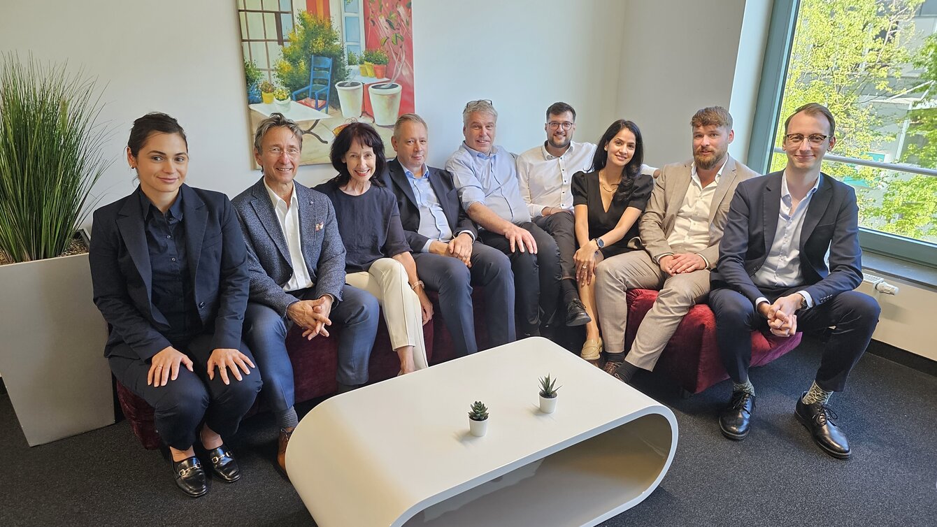 Mehrere Menschen sitzen eng nebeneinander auf einer roten Couch. Im Hintergrund ist ein buntes Bild zusehen, sowie ein Fenster ins Grüne. 