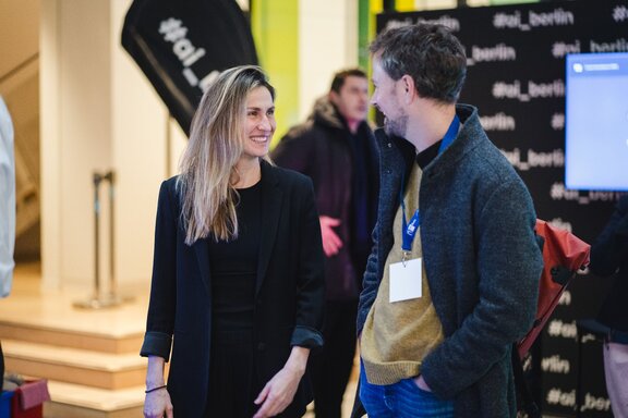 Zu sehen ist eine junge blonde Frau, welche ein schwarzes Sakko trägt und freundlich schaut. Sie unterhält sich stehend mit einem Mann, der eine dunkle Jacke trägt, darunter einen hellbraunen Pullover. Sie befinden sich auf der Digital-Konferenz der IHK Berlin und HTW Berlin.