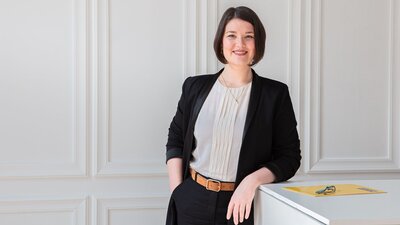 Zu sehen ist eine Frau mit mittellangen dunklen Haaren. Sie trägt eine dunkle Stoffhose, eine helle Bluse und ein dunkles Sakko. Sie lehnt sich an einen Tisch und schaut freundlich in die Kamera.