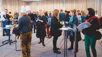 zu sehen sind viele Personen in einem geschlossenen Raum, welche sich an Stehtischen oder frei im Raum mit anderen Anwesenden unterhalten, es herrscht reges Treiben.