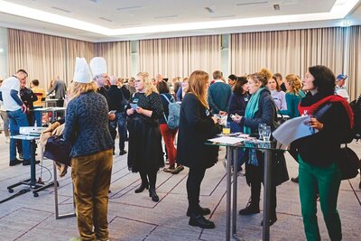zu sehen sind viele Personen in einem geschlossenen Raum, welche sich an Stehtischen oder frei im Raum mit anderen Anwesenden unterhalten, es herrscht reges Treiben.