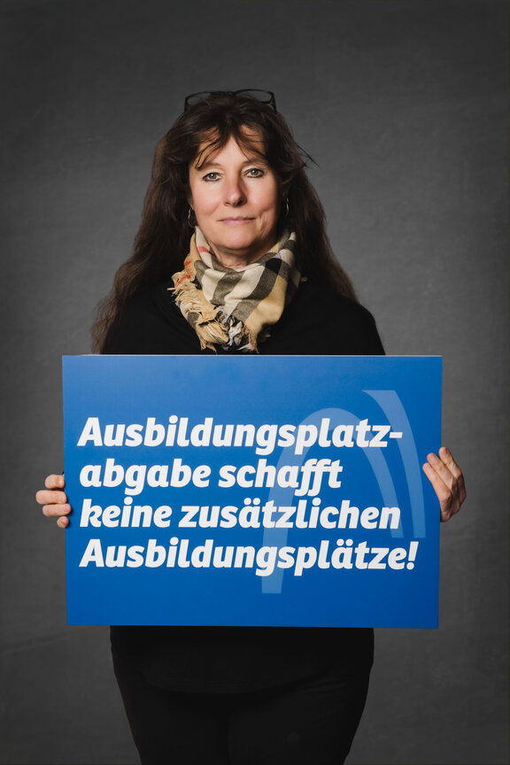 Eine Person steht vor einem neutralen grauen Hintergrund und hält ein großes blaues Schild mit der weißen Aufschrift: "Ausbildungsplatzabgabe schafft keine zusätzlichen Ausbildungsplätze!"