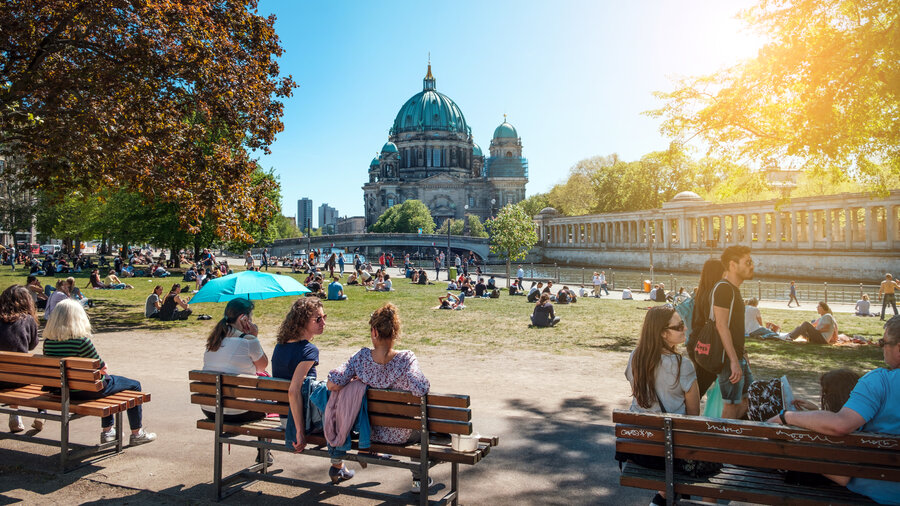 Im Vordergrund befinden sich drei Bänke auf denen Personen sitzen, dahinter liegt eine Wiese. Im Hintergrund befindet sich ein großes Gebäude mit Kuppel - es ist der Berliner Dom.