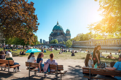 Im Vordergrund befinden sich drei Bänke auf denen Personen sitzen, dahinter liegt eine Wiese. Im Hintergrund befindet sich ein großes Gebäude mit Kuppel - es ist der Berliner Dom.