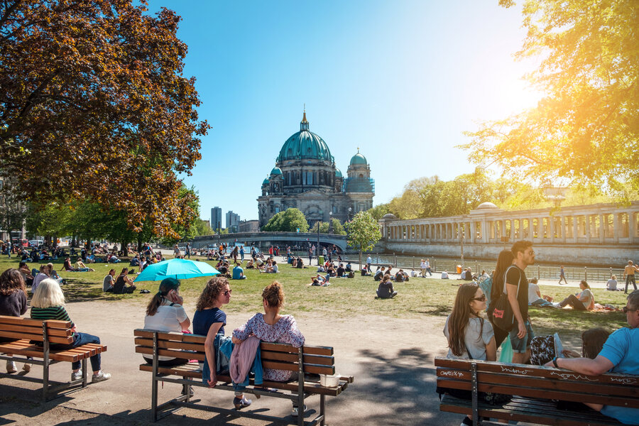 Im Vordergrund befinden sich drei Bänke auf denen Personen sitzen, dahinter liegt eine Wiese. Im Hintergrund befindet sich ein großes Gebäude mit Kuppel - es ist der Berliner Dom.