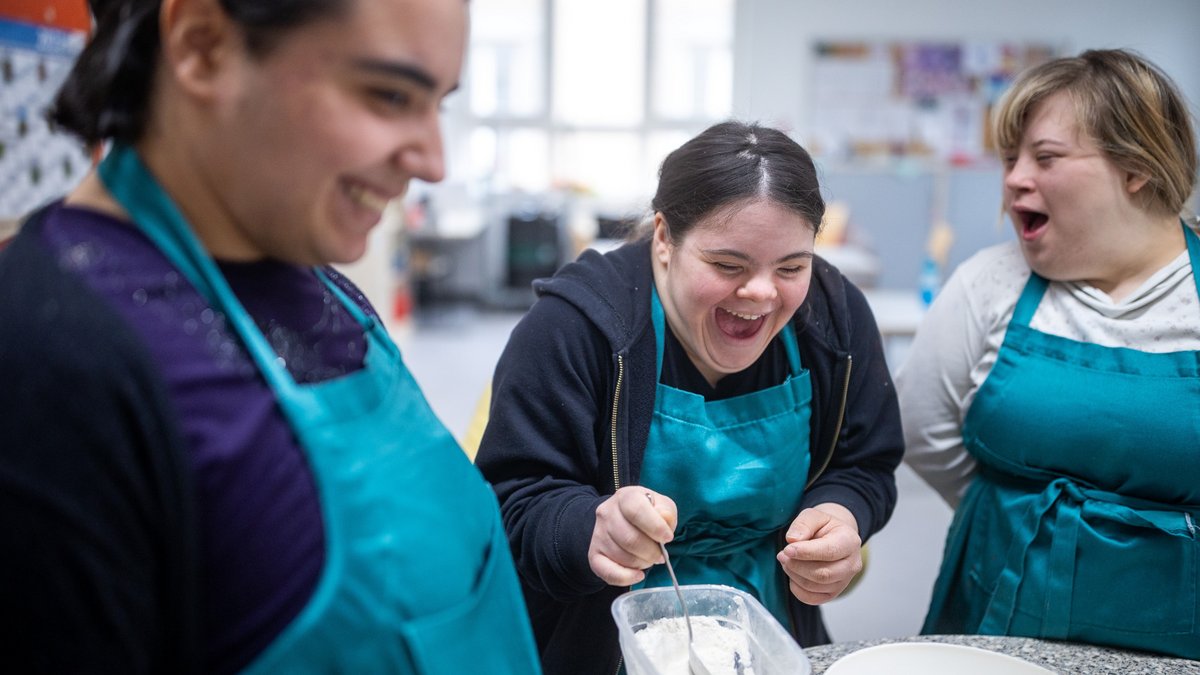 Drei Personen mit türkisfarbenen Schürzen stehen um einen Tisch, eine Person rührt in einem Behälter mit Mehl, auf dem Tisch stehen weitere Schüsseln.