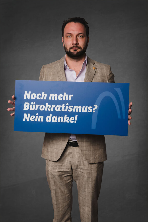 Eine Person steht vor einem neutralen grauen Hintergrund und hält ein großes blaues Schild mit der weißen Aufschrift: "Noch mehr Bürokratismus? Nein danke!"