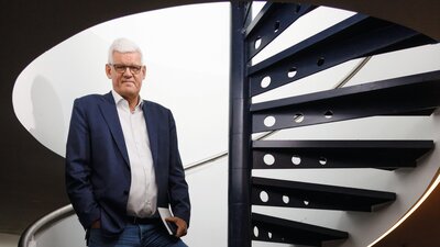 Zu sehen ist ein älterer Mann im Sakko, der in einem Treppenaufgang steht. In einer Hand hält er ein Notizbuch.