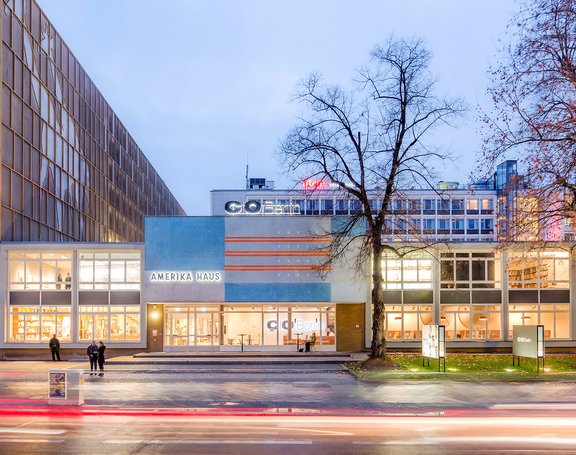 Außenansicht des Amerika Hauses in Berlin bei Nacht mit beleuchteten Fenstern, zwei Bäumen und vorbeifahrendem Verkehr