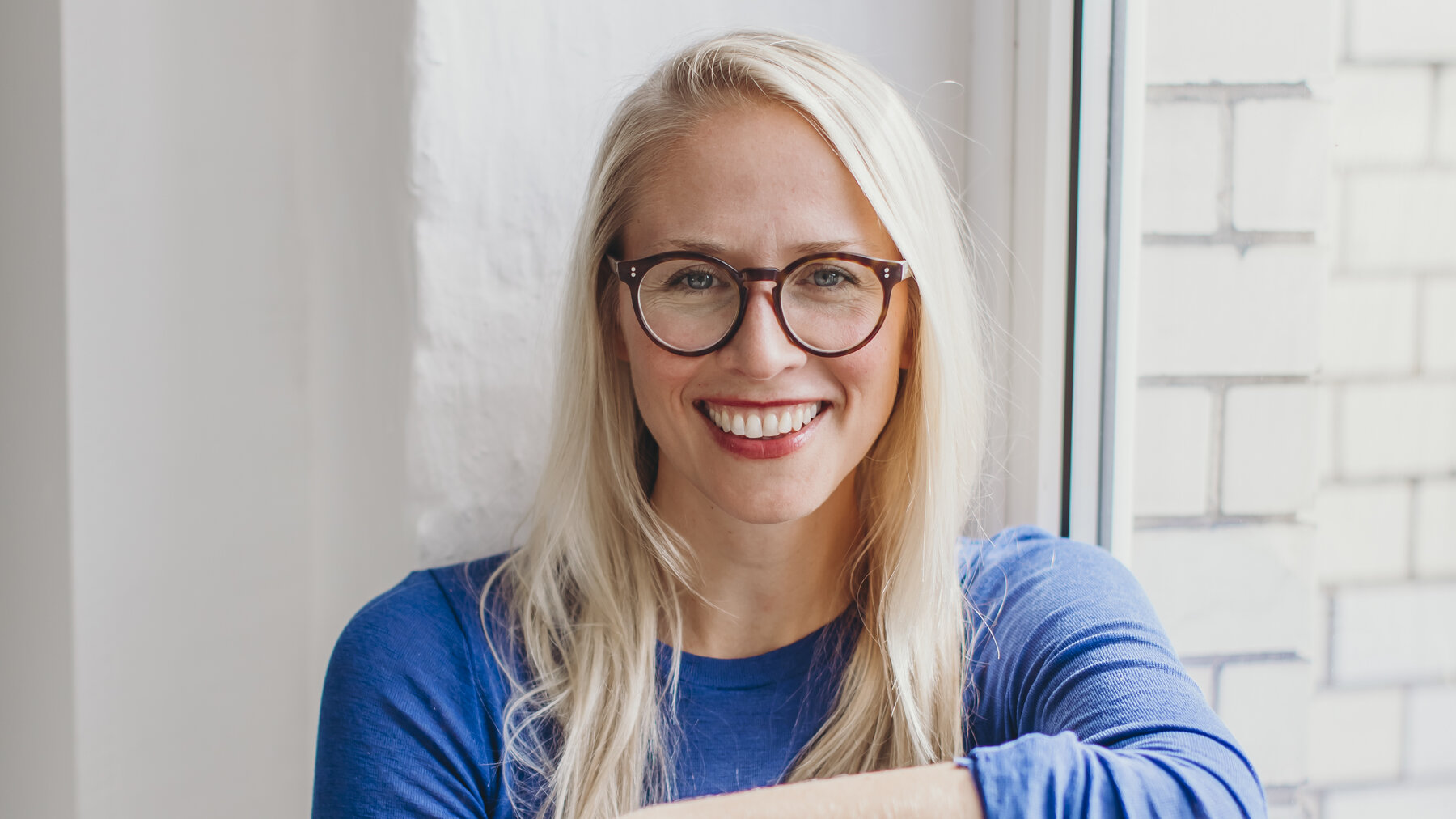 Eine junge, blonde, lachende Frau mit Brille und blauem Oberteil sitzt auf einer Fensterbank. Es ist Susann Hoffmann, Co-Founderin von Edition F und Manti Manti.