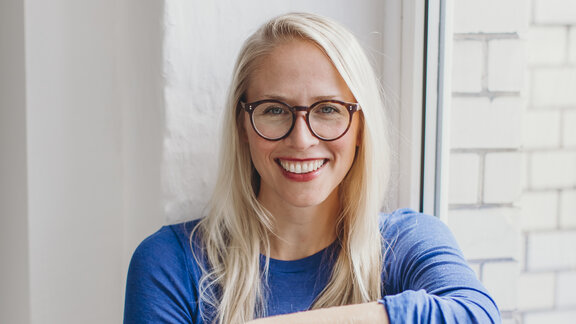 Eine junge, blonde, lachende Frau mit Brille und blauem Oberteil sitzt auf einer Fensterbank. Es ist Susann Hoffmann, Co-Founderin von Edition F und Manti Manti.