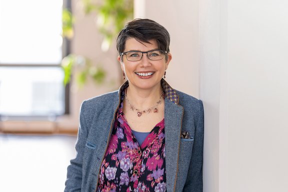 Person mit kurzem, dunkelbraunem Haar trägt blaues Sakko und bunt gemustertes Hemd, lehnt an weißer Wand.