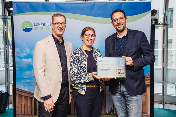 Drei Personen stehen vor einem Banner mit dem Schriftzug 'Klimaschutz Berlin', zwei von ihnen halten gemeinsam eine Urkunde mit dem Titel 'Unser Markt Berlin'