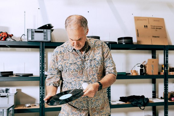 Mann mit gemustertem Hemd hält eine schwarze Vinyl-Schallplatte in einem Lagerraum mit Regalen voller Schallplatten und Kartons