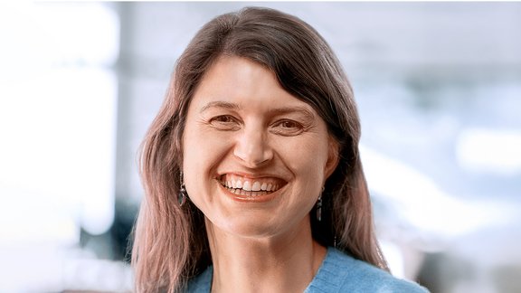 Frau mit langen, gewellten braunen Haaren trägt hellblaues Oberteil und silberne Ohrringe vor unscharfem Hintergrund.