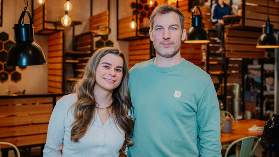 Die Betreiberin und der Betreiber von Fendricks Coffee posieren Arm in Arm in ihrem Café, das sich stylisch im Holz-Look über zwei Etagen erstreckt.