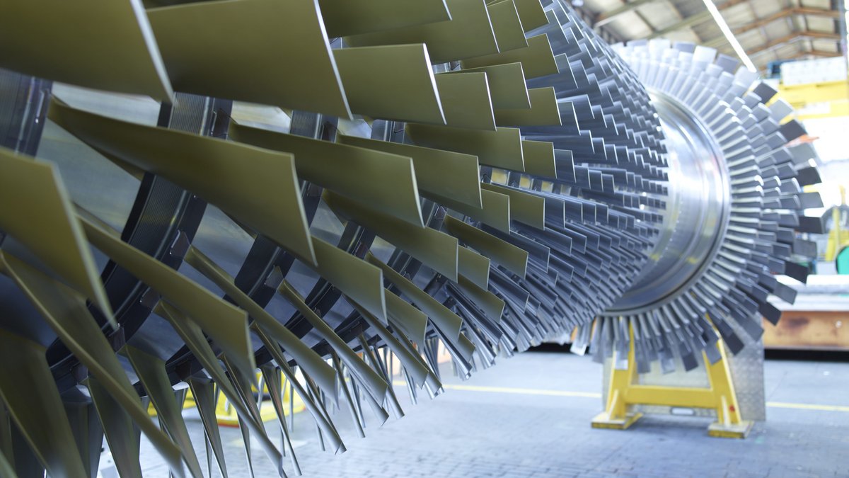 Nahaufnahme einer großen Turbinenwelle mit vielen schräg angeordneten Metallflügeln in einer Industriehalle