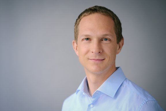 Mann mit kurzem braunem Haar trägt hellblaues Hemd vor grauem Hintergrund.