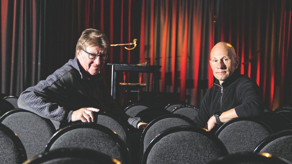 Zwei Männer mittleren Alters sitzen auf Stühlen in einem Theater.