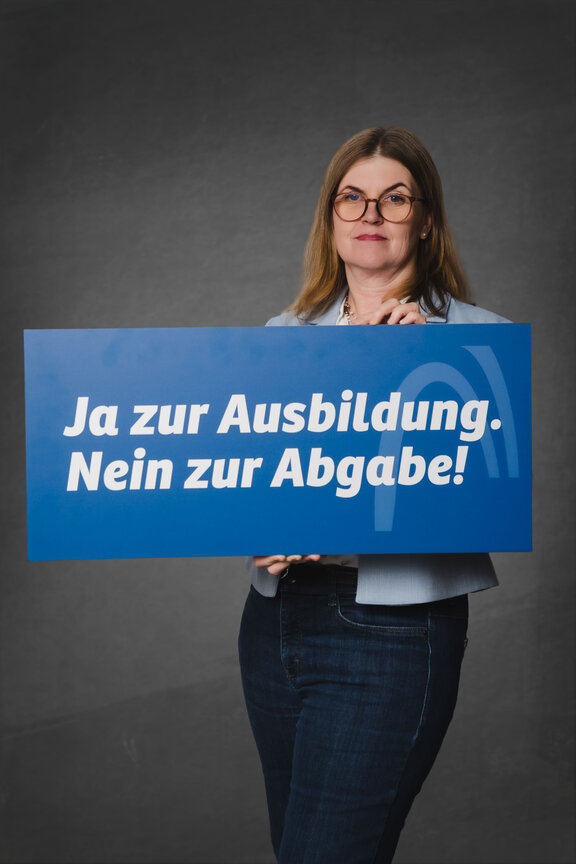 Eine Person steht vor einem neutralen grauen Hintergrund und hält ein großes blaues Schild mit der weißen Aufschrift: "Stoppt die Pläne zur Ausbildungsplatzabgabe!"