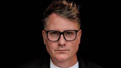 Porträt-Foto eines Mannes mit Brille vor schwarzem Hintergrund. Es ist Scott Lipinski, CEO der Fashion Council Germany.