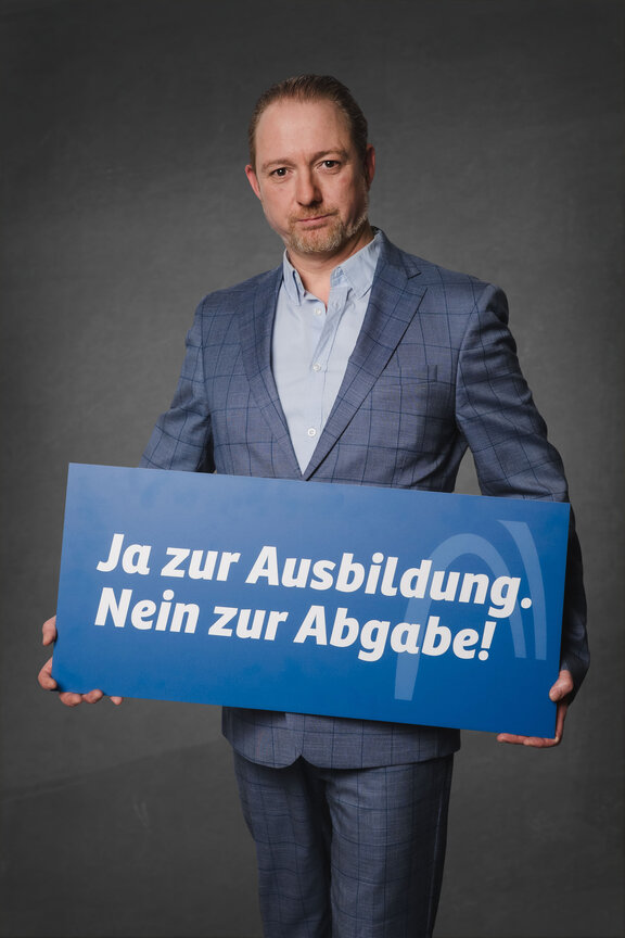 Eine Person steht vor einem neutralen grauen Hintergrund und hält ein großes blaues Schild mit der weißen Aufschrift: "Ja zur Ausbildung. Nein zur Abgabe!"