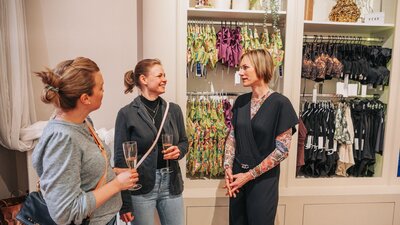 Auf dem Bild sind drei Frauen zu sehen, die sich in einem Verkaufsgespräch zu befinden scheinen. Zwei von ihnen halten ein Sektglas und sie stehen in einem Dessousgeschäft.