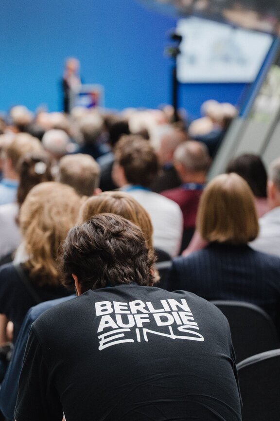 Zu sehen ist das bestuhlte Publikum einer Veranstaltung. Eine Person trägt ein Shirt mit der Aufschrift "Berlin auf die Eins".