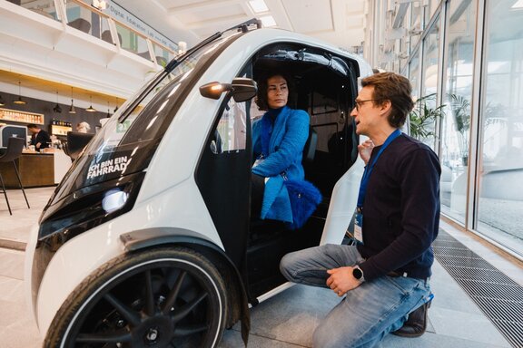 Eine Frau sitzt in einem futuristischen Kleinwagen und unterhält sich mit einem vor der offenen Tür knienden Mann.