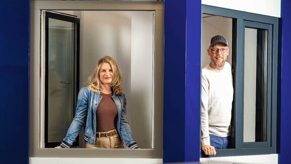 Zu sehen sind ein Mann und eine Frau, die aus je einem Fenster eines Gebäudes freundlich in die Kamera schauen. 