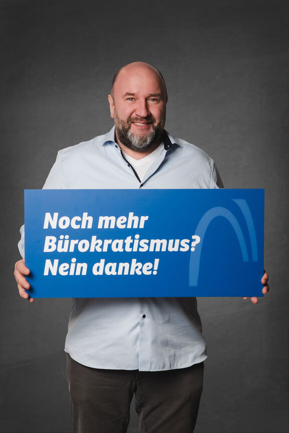 Eine Person steht vor einem neutralen grauen Hintergrund und hält ein großes blaues Schild mit der weißen Aufschrift: "Noch mehr Bürokratismus? Nein danke!"
