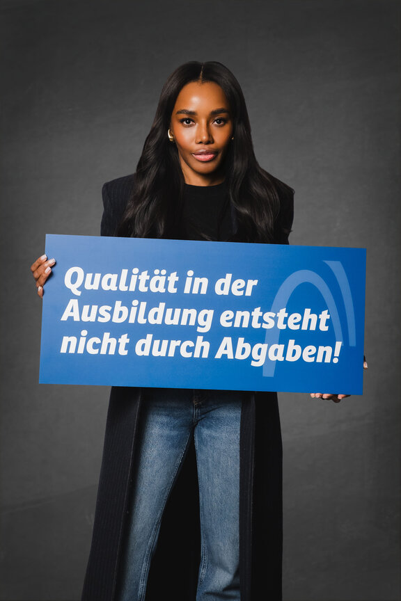 Eine Person steht vor einem neutralen grauen Hintergrund und hält ein großes blaues Schild mit der weißen Aufschrift: "Qualität in der Ausbildung entsteht nicht durch Abgaben!"