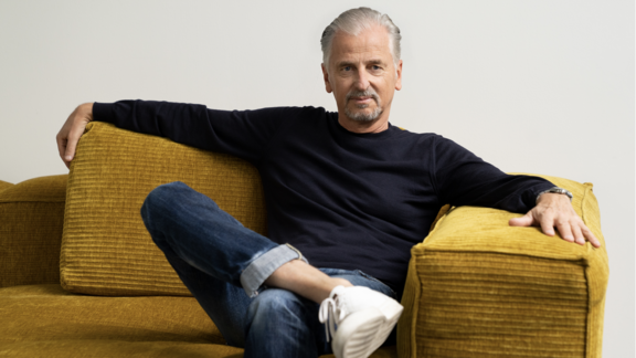 Mann sitzt entspannt auf einem senfgelben Cordsofa, trägt dunkles Langarmshirt, blaue Jeans und weiße Sneaker.