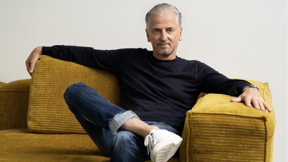 Mann sitzt entspannt auf einem senfgelben Cordsofa, trägt dunkles Langarmshirt, blaue Jeans und weiße Sneaker.