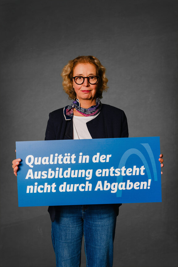 Eine Person steht vor einem neutralen grauen Hintergrund und hält ein großes blaues Schild mit der weißen Aufschrift: "Qualität in der Ausbildung entsteht nicht durch Abgabe!"