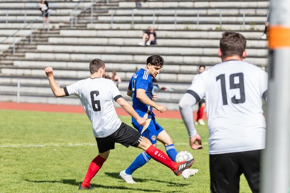 Fußballspieler in blauer und weißer Mannschaftskleidung kämpfen um den Ball auf dem Spielfeld vor leeren Tribünen