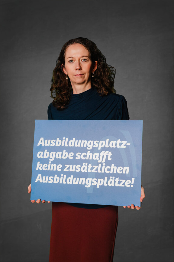 Eine Person steht vor einem neutralen grauen Hintergrund und hält ein großes blaues Schild mit der weißen Aufschrift: "Ausbildungsplatzabgabe schafft keine weiteren Ausbildungsplätze!"