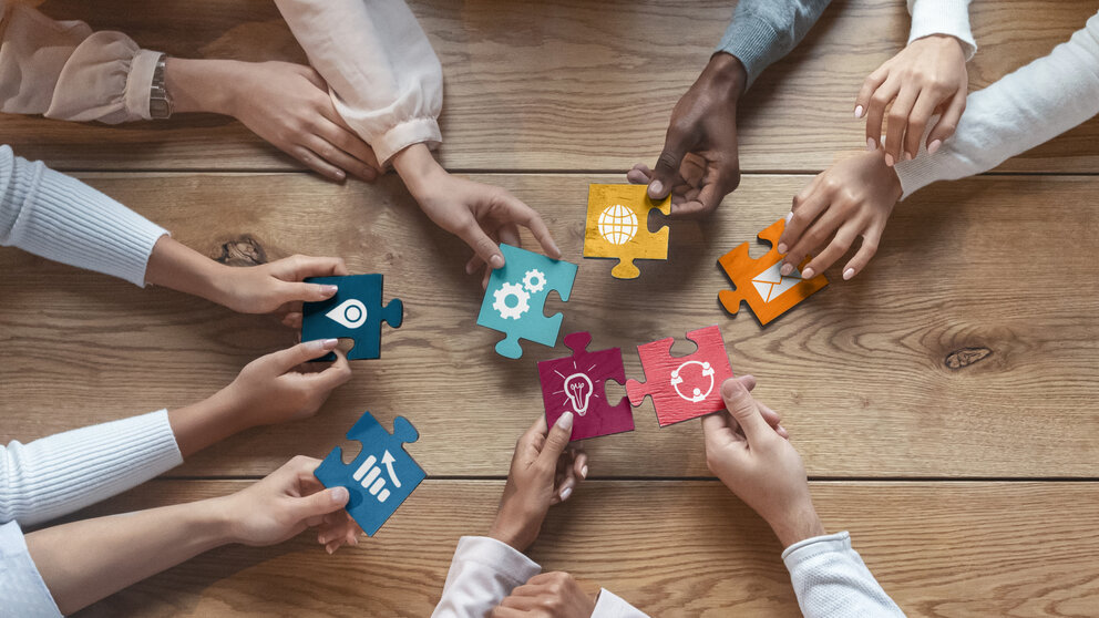 Zu sehen ist eine hölerne Tischpatte von oben. Man sieht mehrere Arme und Hände, ohne den restlichen Körper der Personen. In den Händen werden bunte Puzzleteile in die Tischmitte gehalten.