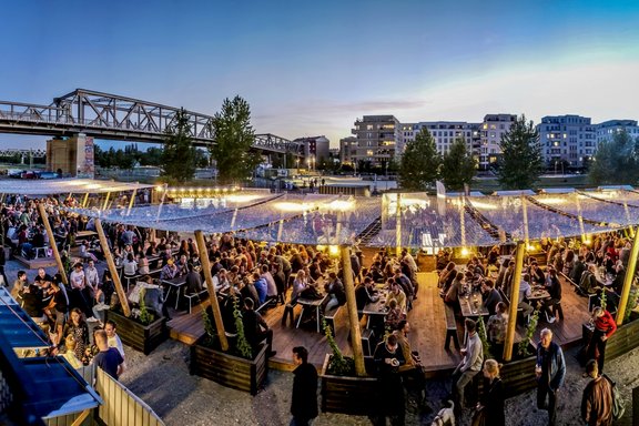 Menschen sitzen und stehen in einem belebten Biergarten am Wasser bei Abenddämmerung, im Hintergrund eine Eisenbahnbrücke und moderne Wohngebäude