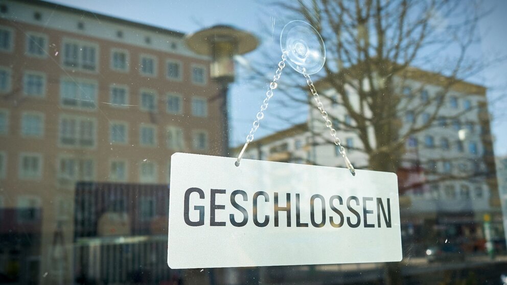 zu sehen ist ein Schaufenster, in dem ein Schild mit der Aufschrift "geschlossen" steht. Im Glas des Fensters spiegeln sich die Gebäude der anderen Straßenseite.