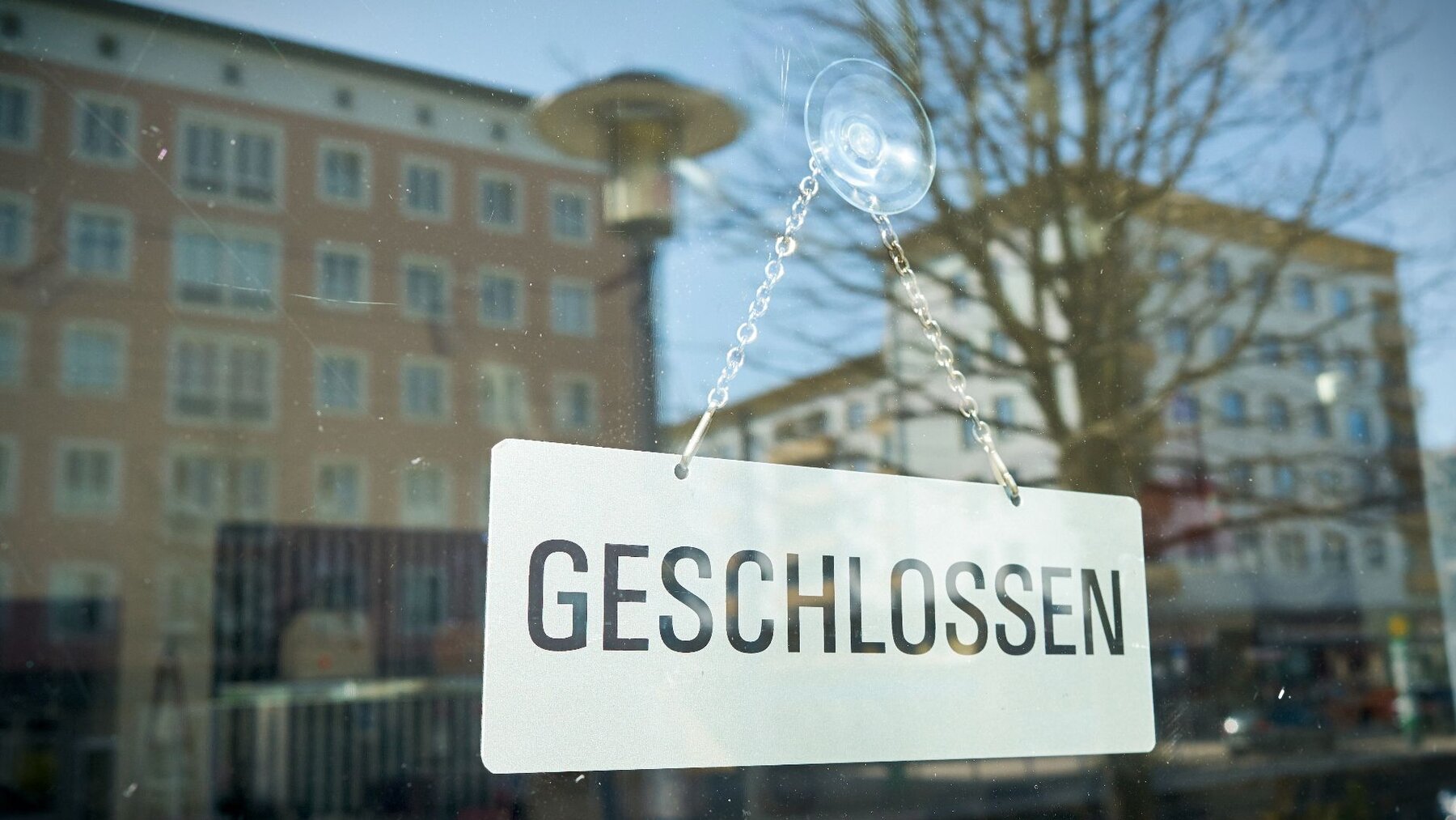 zu sehen ist ein Schaufenster, in dem ein Schild mit der Aufschrift "geschlossen" steht. Im Glas des Fensters spiegeln sich die Gebäude der anderen Straßenseite.