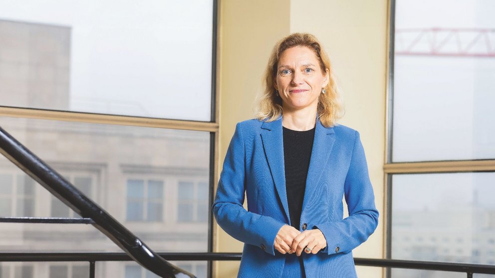 Eine blonde Frau mittleren Alters in einem blauen Hosenanzug steht vor einem Fenster.