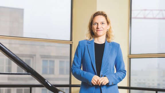 Eine blonde Frau mittleren Alters in einem blauen Hosenanzug steht vor einem Fenster.