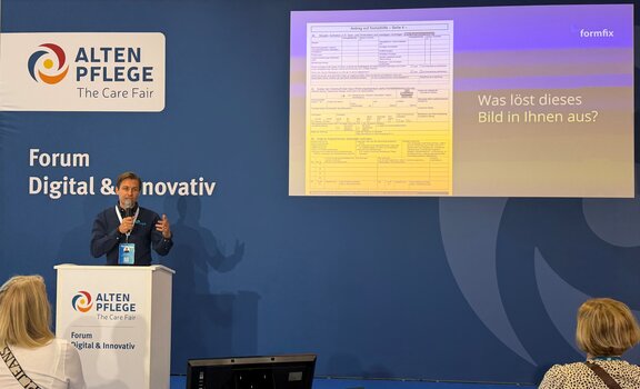 Ein Mann steht hinter einem weißen Rednerpult und hinter ihm ist eine blaue Wand mit einer Präsentation 
