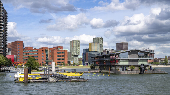 Ein Holzgebäude schwimmt auf dem Wasser neben einem Bootssteg. Im Hintergrund stehen Hochhäuser.