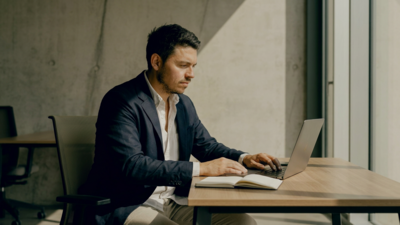 Ein Mann im Anzug sitzt vor seinem Laptop und guckt auf den Bildschirm. Hinter ihm ist eine betonfarbene Wand zusehen.